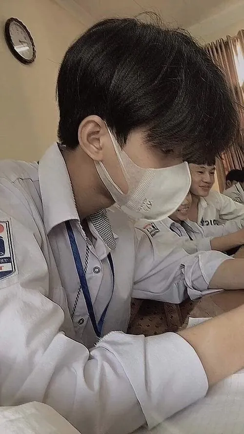 Ảnh nam sinh lớp 7 giấu mặt với khẩu trang trắng, mặc đồng phục và đeo dây thẻ trong lớp học có bạn ngồi phía sau.