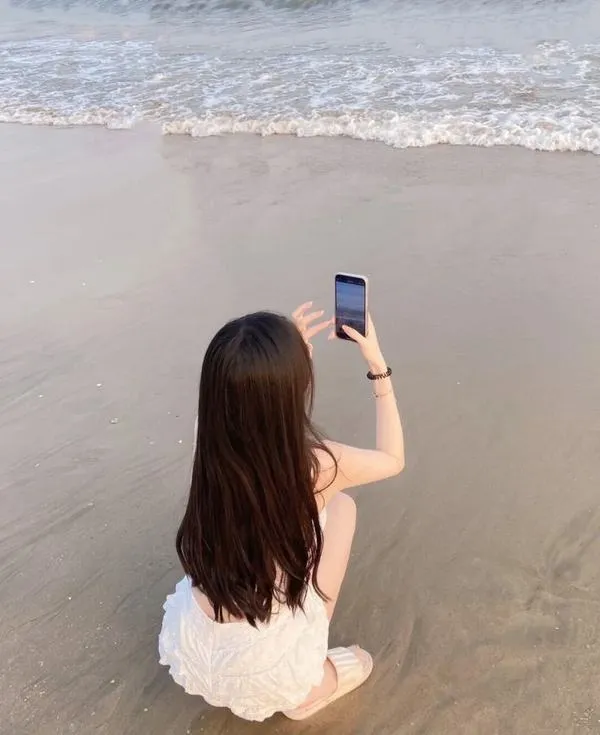 Nàng ngồi sát nước, cầm điện thoại chụp ảnh, tóc thả lưng, vẻ kín đáo dễ thương.