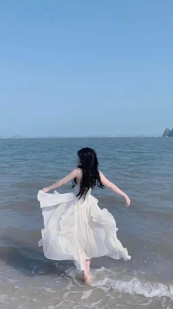 Cô gái bước nhanh trên bờ cát, tà váy tung nhẹ, dáng lưng thon, vibe mùa hè.