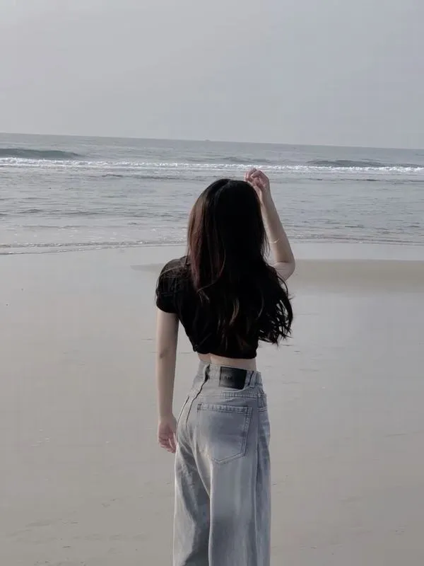 Cô gái quay lưng trên bãi biển, tóc dài bay nhẹ, dáng đứng mảnh, mood yên ả.