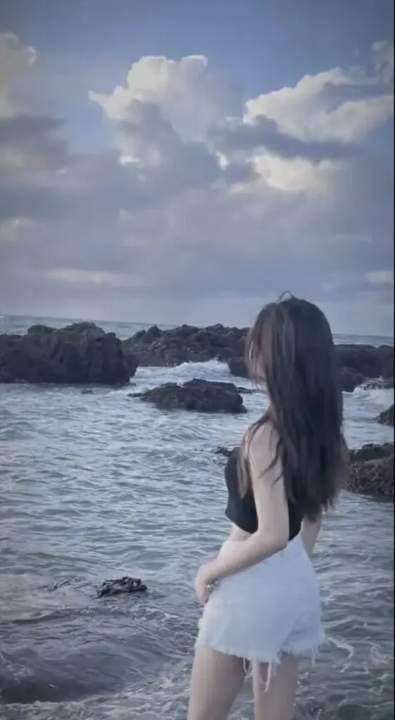 Cô gái nhìn ra xa trên bờ biển, váy trắng gọn, tóc bay, nét yên bình dịu nhẹ.