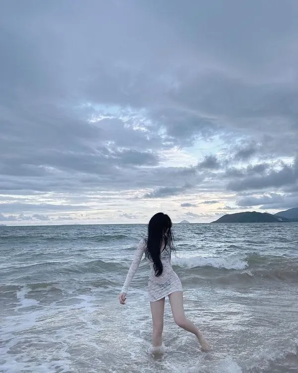 Kính râm che mắt, cô gái đứng sát nước, tóc bay, nét lạnh lùng đầy cuốn hút.