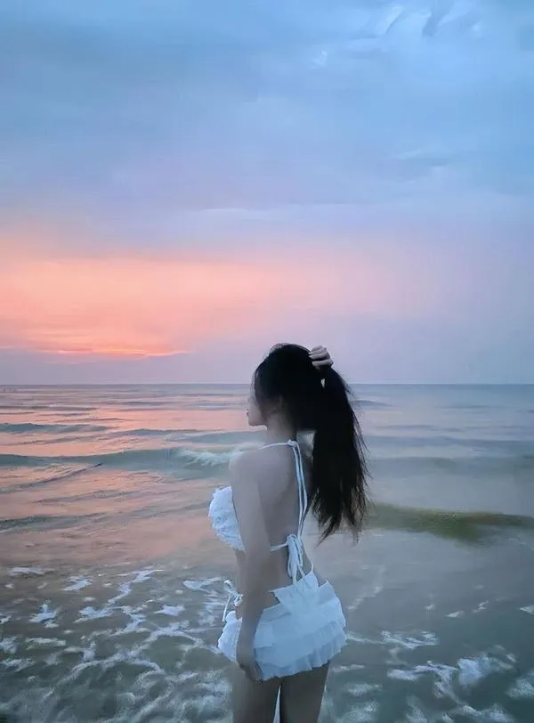 Nàng bước sát sóng biển lúc hoàng hôn, tóc đen ướt nhẹ, dáng nghiêng gợi tò mò.