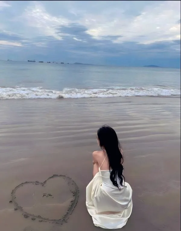 Cô gái ngồi trên cát trắng vẽ trái tim, tóc phủ lưng, ánh nhìn giấu kín sau vai.