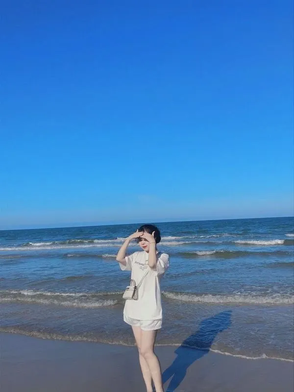 Nàng mặc đồ sáng màu, đứng dưới ánh nắng, một tay che mặt, đường nét dịu dàng.
