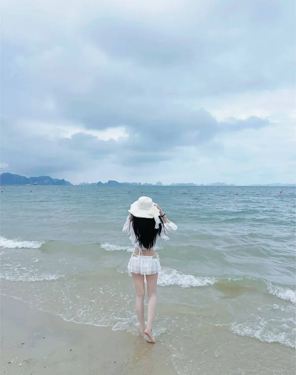 Cô gái thả bước ở mép nước, mũ rộng vành che nửa mặt, dáng nhỏ nhắn thanh thoát.