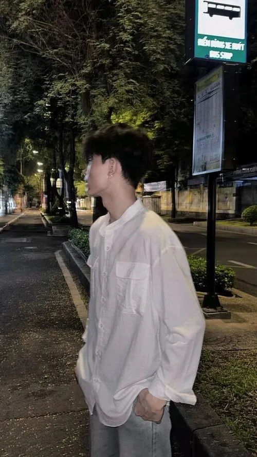 Hình ảnh hotboy sơ mi trắng nhìn nghiêng trong khung cảnh tối, ánh sáng yếu làm bóng mặt và cổ áo hiện nhẹ.